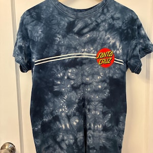 May include: A short-sleeve crew neck t-shirt with a dark blue and white tie-dye pattern. The front features two horizontal white lines across the chest and a circular red and yellow logo with the text SANTA CRUZ printed in black lettering.