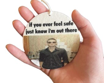 Op de afbeelding: Ronde witte autoluchtverfrisser met een foto van een man met een zonnebril en de tekst "if you ever feel safe just know i'm out there".