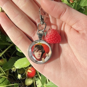 May include: A silver keychain with a round glass charm featuring a photo of a person with brown hair and a red strawberry. The charm is attached to a silver chain with a silver ring.