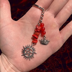 May include: A silver keychain with a red stone charm, a silver crown charm, and a silver charm with a pentagram inside a sun.