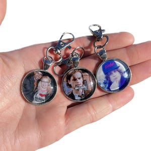 May include: Three silver keychains with round glass charms. The charms feature images of people. One charm has a photo of a man with the text "actually" printed below. Another charm has a photo of a man and woman. The third charm has a photo of a person wearing a blue hat with the text "slay girl" printed on it.