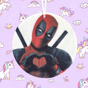 Puede incluir: Un adorno redondo y blanco con una imagen detallada de Deadpool con su traje rojo y negro característico, formando un corazón con sus manos. El adorno tiene una cuerda blanca para colgar. El fondo es de color lila claro con patrones de unicornios y arcoíris.