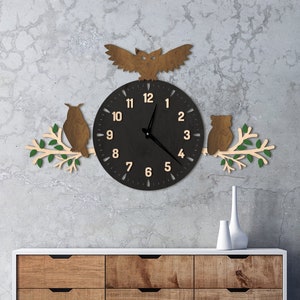 Peut inclure: Une horloge murale en bois sur le thème des hiboux avec un cadran noir et des chiffres clairs. Deux hiboux sont perchés sur des branches avec des feuilles vertes, encadrant l'horloge. Un troisième hibou est assis au-dessus de l'horloge. L'horloge est fixée sur un mur gris au-dessus d'une commode en bois.