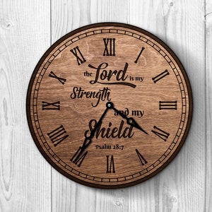 Puede incluir: Un reloj de madera redondo con un marco marrón oscuro y números romanos. La esfera del reloj presenta el texto "the Lord is my Strength and my Shield" y "psalm 28:7". Las manecillas del reloj señalan aproximadamente las 4:00.