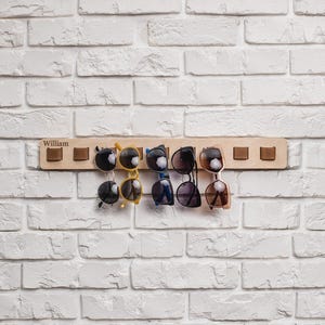 A wooden wall-mounted sunglasses holder labeled “William” holds six pairs of sunglasses by leather loops. The holder is attached to a white-painted brick wall and has eight evenly spaced loops, with two empty on each end. The sunglasses vary in shape and color, including black, yellow, blue, and brown frames.