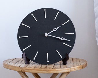 May include: A modern black wall clock with white stick hour markers and hands. The clock is displayed on a small bamboo side table. The clock's design is minimalist and contemporary, suitable for home decor.