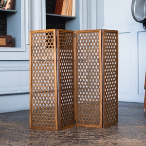 May include: A three-panel room divider made of wood, featuring a woven circular design. The divider is a warm brown colour and stands on a dark wood floor. The background includes a bookshelf, a vintage lamp, and a light blue wall.