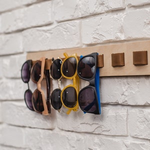 May include: A wooden sunglasses rack with several pairs of sunglasses hanging from it. The sunglasses are various colors, including black, yellow, and brown. The rack is mounted on a white brick wall. The rack has brown leather straps.