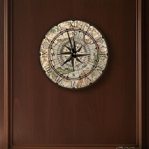 May include: A round wall clock with a compass rose design. The clock face features a map-like background with Roman numerals and a compass needle. The clock is mounted on a dark brown wooden wall.
