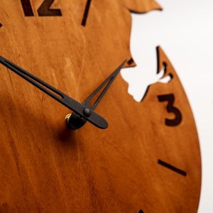 May include: Close-up of a wooden wall clock with a natural wood grain finish. The clock features black hands and black numerals. The design includes a cutout shape, adding a unique aesthetic to the timepiece. The clock's hands point to approximately 12:15.