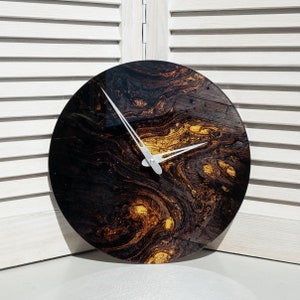 Op de afbeelding: Ronde wandklok met een donkerbruin en goud wervelend patroon. De klok heeft zilveren wijzers en een houtnerf-look. Een decoratief item voor in huis.