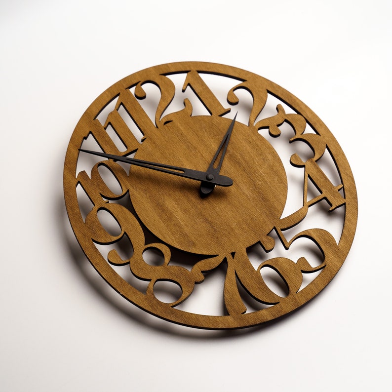 Puede incluir: Un reloj de madera redondo con acabado de madera natural. La esfera del reloj presenta n&uacute;meros recortados y un c&iacute;rculo central. Las manecillas negras indican la hora. Es un reloj de pared decorativo.