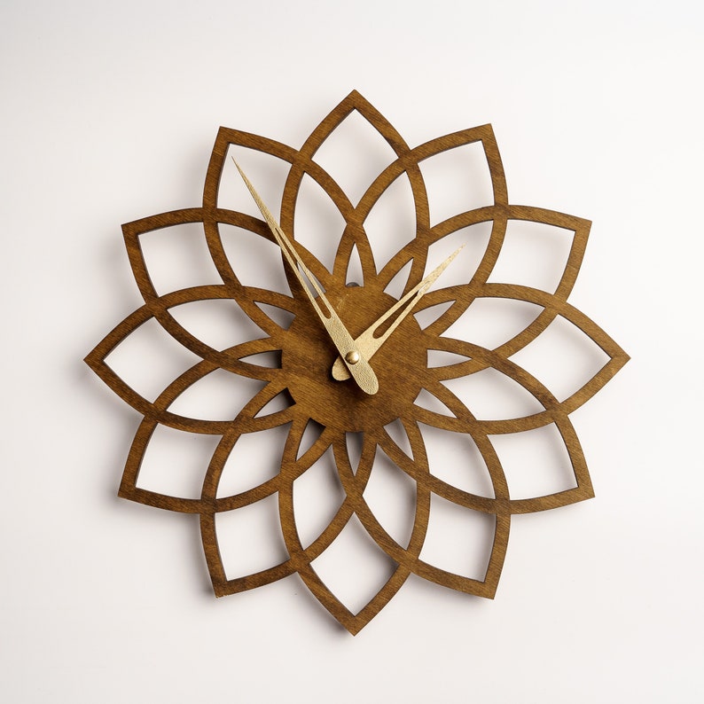 Puede incluir: Un reloj de pared de madera con un dise&ntilde;o floral. La esfera del reloj es de color marr&oacute;n oscuro, con un patr&oacute;n recortado que se asemeja a una flor. Las manecillas del reloj son de color dorado. El reloj est&aacute; montado en una pared blanca.