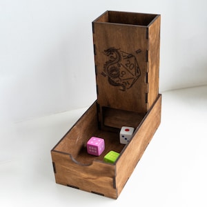 Può includere: Un set di torre e vassoio per dadi in legno. La torre presenta un drago e un design di dado D20. Tre dadi multicolori sono nel vassoio. Il legno ha una calda macchia marrone. Il set è progettato per i giochi da tavolo.