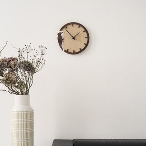 May include: A round wooden wall clock with a light wood face and a dark brown frame. A dark brown bird silhouette is cut out of the frame. The clock hands are black. The clock is mounted on a white wall.