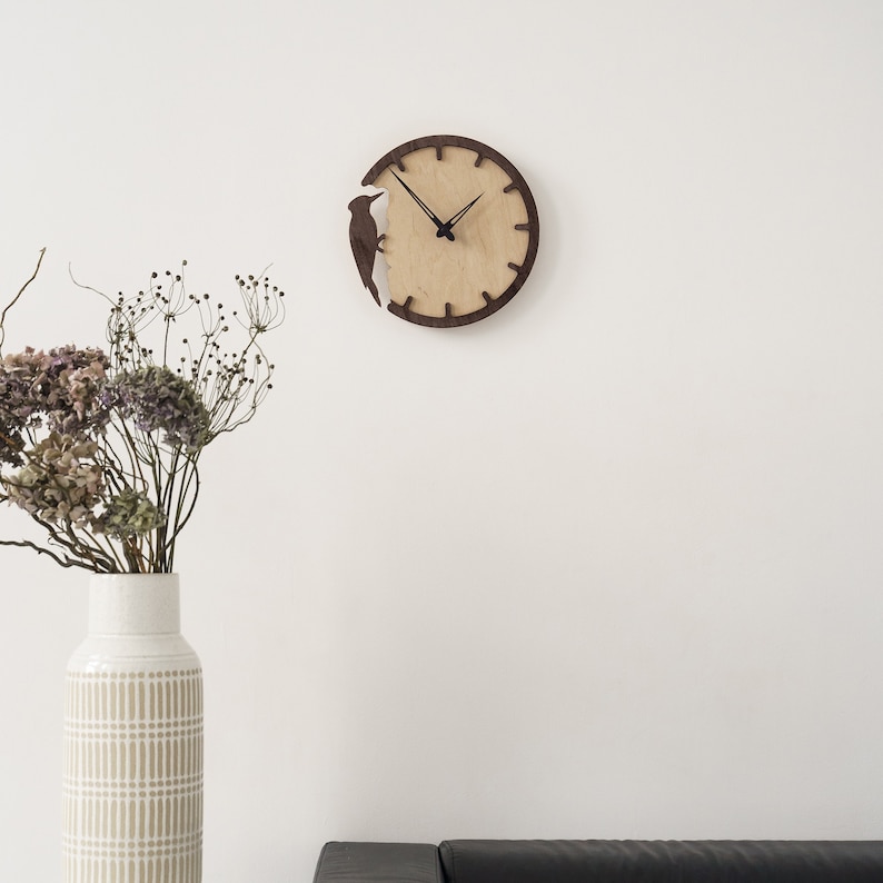 May include: A round wooden wall clock with a light wood face and a dark brown frame. A dark brown bird silhouette is cut out of the frame. The clock hands are black. The clock is mounted on a white wall.