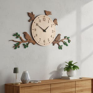 Op de afbeelding: Een ronde houten wandklok met zwarte wijzers en cijfers van 1 tot 12. De klok is voorzien van decoratieve bruine houten vogelsilhouetten en takken met kleine groene blaadjes die aan de zijkanten uitsteken, gemonteerd op een lichte muur.