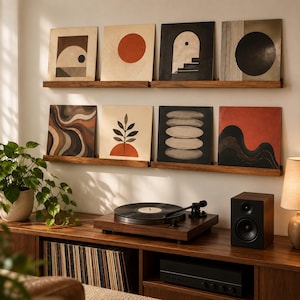 May include: Two wooden wall shelves display eight square art prints featuring abstract geometric shapes, wavy lines, a plant motif, and stacked stones in shades of black, white, brown, and orange, positioned above a turntable and speaker.