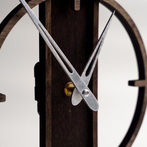 May include: Close-up of a modern wooden clock with a dark brown finish. The clock features silver-colored hands and a circular design. The clock's hands are positioned to indicate the time. The clock is mounted on a dark brown wooden base.
