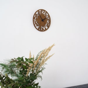 Puede incluir: Reloj de pared ovalado de madera con n&uacute;meros recortados y manecillas negras. El reloj est&aacute; montado en una pared blanca. La esfera del reloj es marr&oacute;n. El reloj est&aacute; encima de una exhibici&oacute;n de vegetaci&oacute;n y hierbas secas.