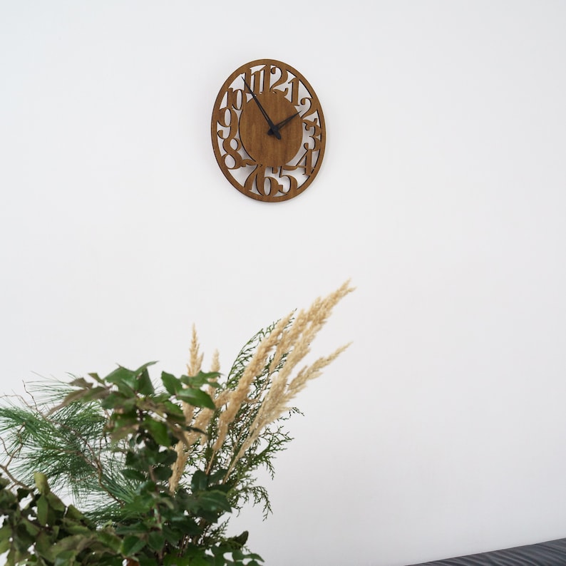 Puede incluir: Reloj de pared ovalado de madera con n&uacute;meros recortados y manecillas negras. El reloj est&aacute; montado en una pared blanca. La esfera del reloj es marr&oacute;n. El reloj est&aacute; encima de una exhibici&oacute;n de vegetaci&oacute;n y hierbas secas.