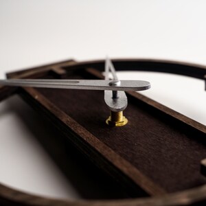 May include: Close-up of a modern wall clock with a dark brown wooden frame and silver hands. The clock's hands are positioned to indicate a specific time. The clock's design is minimalist and contemporary.