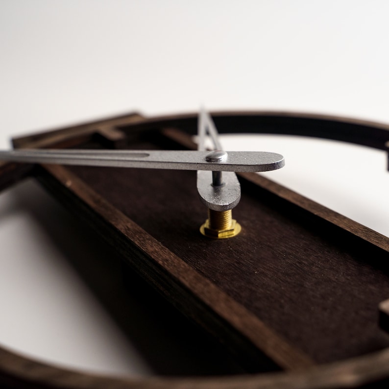 May include: Close-up of a modern wall clock with a dark brown wooden frame and silver hands. The clock's hands are positioned to indicate a specific time. The clock's design is minimalist and contemporary.