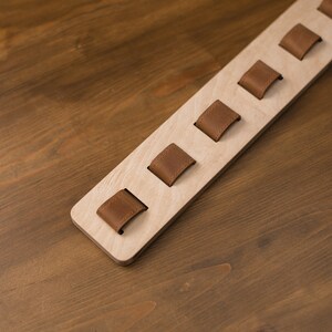 May include: A light-colored wooden organizer with six brown leather loops. The loops are evenly spaced along the length of the wood. The organizer is designed to hold items and is set against a wooden surface.