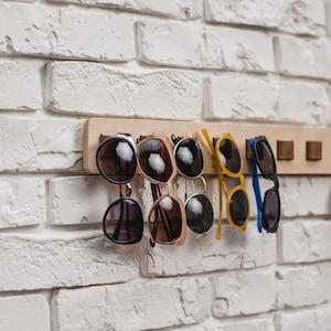 May include: A wooden sunglasses rack with several pairs of sunglasses hanging from it. The sunglasses have various frame colors, including black, brown, and yellow. The rack is mounted on a white brick wall.