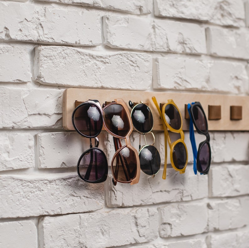 May include: A wooden sunglasses rack with several pairs of sunglasses hanging from it. The sunglasses have various frame colors, including black, brown, and yellow. The rack is mounted on a white brick wall.