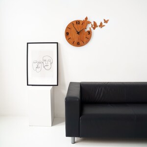May include: A wooden wall clock with a butterfly design. The clock face is brown with black numbers and hands. Butterflies appear to be emerging from the clock. A framed line art print and a black leather sofa are also in the image.