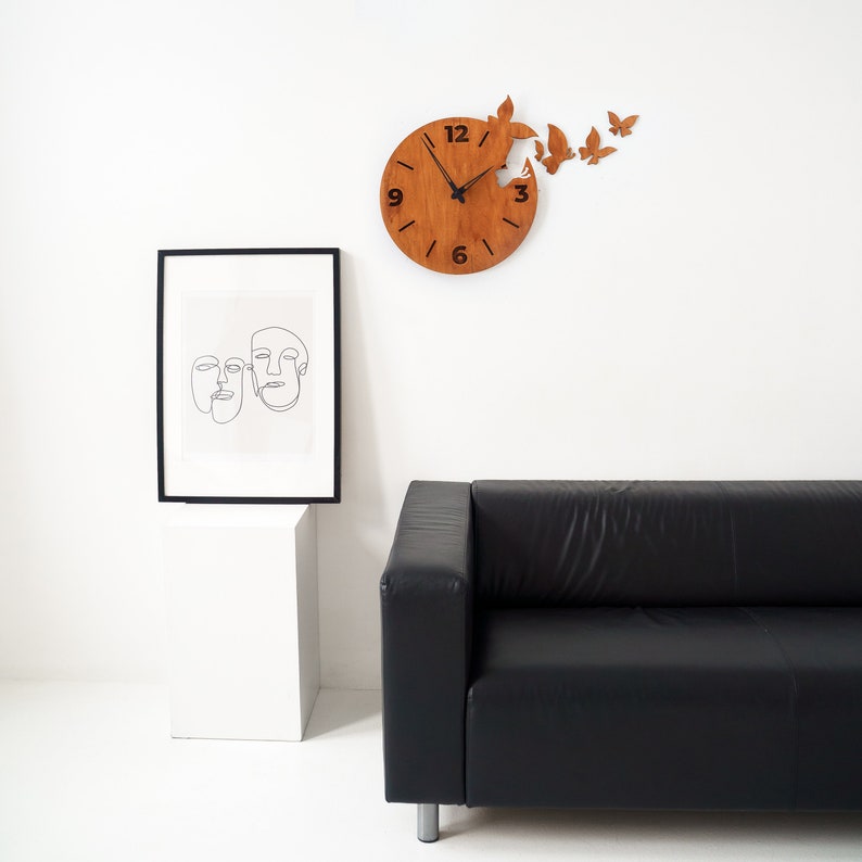 May include: A wooden wall clock with a butterfly design. The clock face is brown with black numbers and hands. Butterflies appear to be emerging from the clock. A framed line art print and a black leather sofa are also in the image.