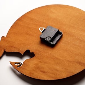May include: A wooden clock face with a cutout design, stained in a warm brown hue. A black clock mechanism is attached to the wooden surface. The clock face has a unique shape, possibly a cat silhouette.