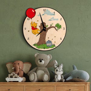 May include: A round wooden wall clock featuring Winnie the Pooh holding a red balloon. The clock face has black hands and numbers, with a tree and honey pot illustration. The clock is surrounded by stuffed animals on a wooden surface.