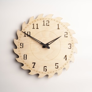 May include: A wooden wall clock with a circular saw blade design. The clock face is light brown with black numerals and hands. The saw blade edge has evenly spaced teeth. The clock measures approximately 12 inches in diameter.