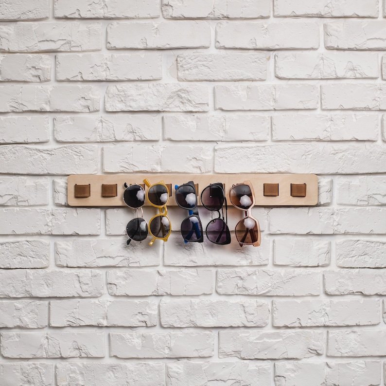 May include: A wooden sunglasses rack with six pairs of sunglasses hanging from it. The rack is mounted on a white brick wall. The sunglasses are various colors, including black, yellow, and brown.