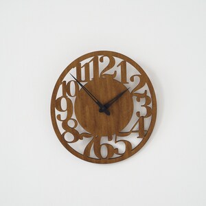 Puede incluir: Un reloj de pared redondo de madera con acabado de madera natural. El reloj presenta n&uacute;meros recortados y manecillas negras. El reloj tiene aproximadamente 30 cm de di&aacute;metro y est&aacute; montado en una pared blanca.