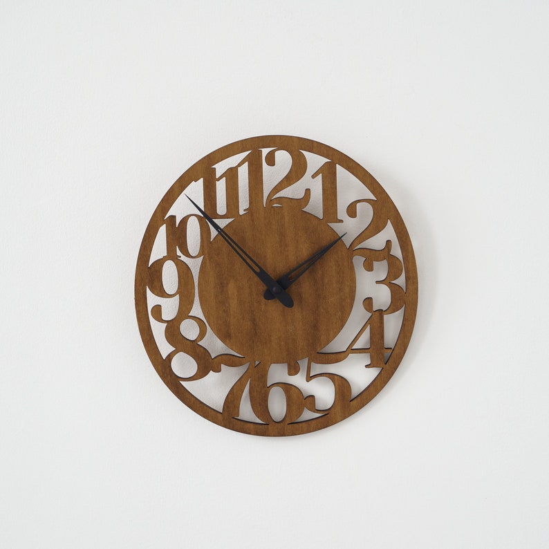 Puede incluir: Un reloj de pared redondo de madera con acabado de madera natural. El reloj presenta n&uacute;meros recortados y manecillas negras. El reloj tiene aproximadamente 30 cm de di&aacute;metro y est&aacute; montado en una pared blanca.
