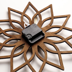 K&ouml;nnte beinhalten: Eine Wanduhr aus Holz mit floralem Design. Die Uhr hat einen dunkelbraunen, lasergeschnittenen Holzrahmen in einem symmetrischen Muster. Das Uhrwerk ist schwarz und auf der R&uuml;ckseite montiert.