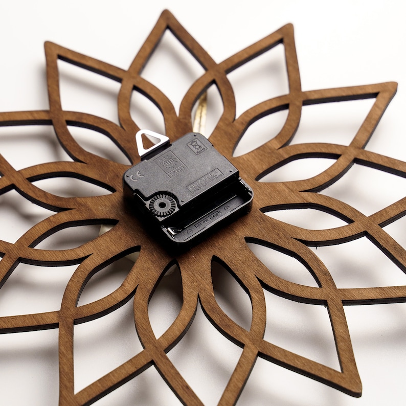 K&ouml;nnte beinhalten: Eine Wanduhr aus Holz mit floralem Design. Die Uhr hat einen dunkelbraunen, lasergeschnittenen Holzrahmen in einem symmetrischen Muster. Das Uhrwerk ist schwarz und auf der R&uuml;ckseite montiert.