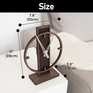 May include: A wooden desk clock with a dark brown finish. The clock has a rectangular base and a vertical support with an oval clock face. The clock is 11 inches (28cm) tall, 7.8 inches (20cm) wide, and 2.9 inches (7.5cm) deep. The word "Size" is at the top.