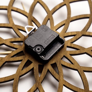 Puede incluir: Primer plano de una esfera de reloj de madera con un dise&ntilde;o floral cortado con l&aacute;ser. El mecanismo del reloj es negro con un colgador de metal plateado. La esfera de madera es de color marr&oacute;n c&aacute;lido y el fondo es blanco.