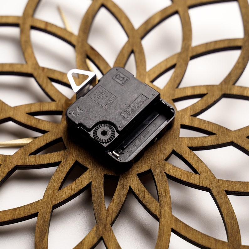 Puede incluir: Primer plano de una esfera de reloj de madera con un dise&ntilde;o floral cortado con l&aacute;ser. El mecanismo del reloj es negro con un colgador de metal plateado. La esfera de madera es de color marr&oacute;n c&aacute;lido y el fondo es blanco.