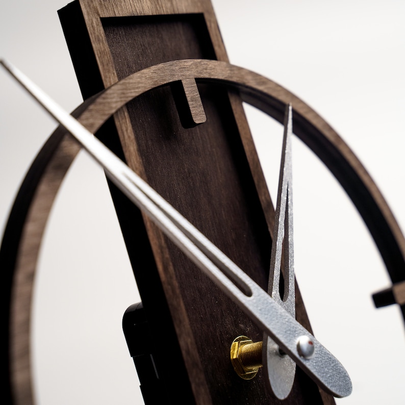 May include: Close-up of a modern wooden clock with a dark brown finish. The clock features a circular design with silver-colored hands and a gold-colored screw. The clock's hands are positioned to indicate the time.