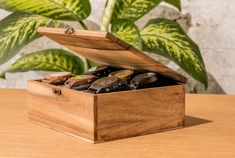 Pode incluir: Uma caixa de rel&oacute;gios de madeira com a tampa ligeiramente aberta, revelando v&aacute;rios rel&oacute;gios com v&aacute;rios designs de pulseira e mostrador. A caixa &eacute; feita de madeira castanha clara e assenta numa superf&iacute;cie de madeira. Os rel&oacute;gios est&atilde;o colocados num forro de veludo preto.