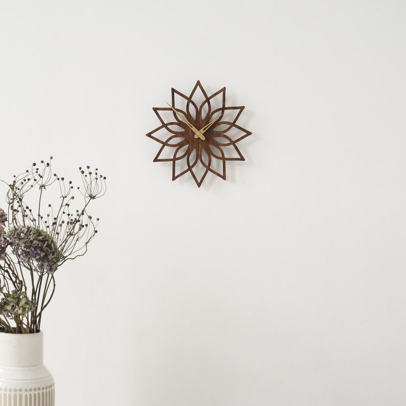 K&ouml;nnte beinhalten: Eine braune Wanduhr aus Holz mit floralem Design, an einer wei&szlig;en Wand befestigt. Die Uhr hat goldfarbene Zeiger. Das Design der Uhr &auml;hnelt einer Blume. Eine Vase mit getrockneten Blumen befindet sich in der linken unteren Ecke. Durchmesser ca. 30 cm.