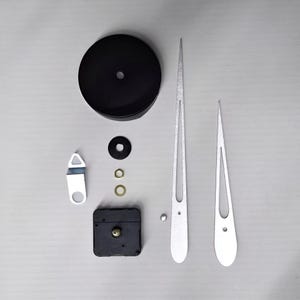May include: Clock making kit with various components. Includes a black clock face cover, silver clock hands, a black clock movement, and other small hardware pieces. The hands are long and slender.