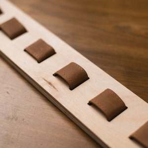 May include: A wooden watch holder with several brown leather watch holders. The watch holder is made of light-colored wood and has a rectangular shape. The leather holders are evenly spaced along the wood.