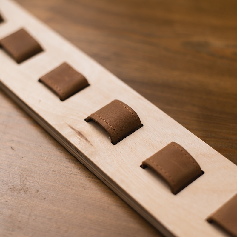 May include: A wooden watch holder with several brown leather watch holders. The watch holder is made of light-colored wood and has a rectangular shape. The leather holders are evenly spaced along the wood.