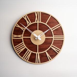 May include: A round wooden wall clock with a dark brown center and a light brown outer ring. The clock features Roman numerals and white clock hands. The clock is a decorative item for home decor.